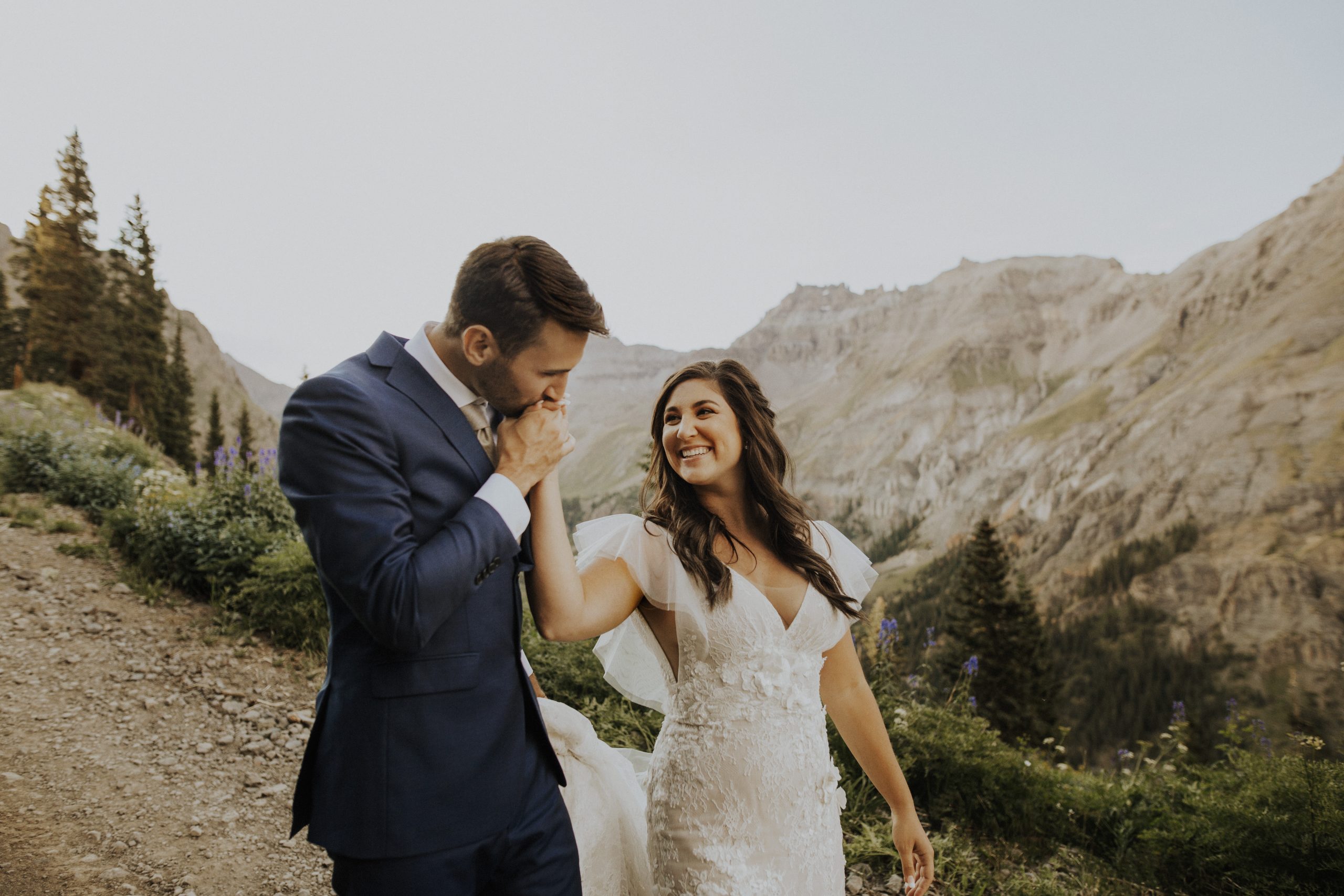 Groom kisses brides hand as they walk down gravel road, with mountains and wildflowers behind them.